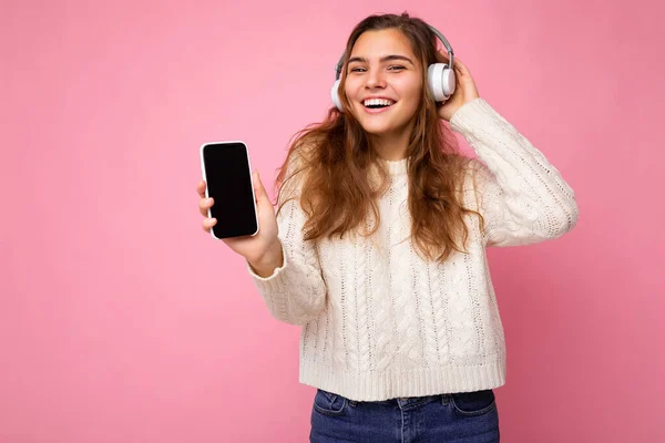 Krásná šťastná usmívající se mladá žena na sobě stylové ležérní oblečení izolované na pozadí zdi držení a zobrazení mobilního telefonu s prázdným displejem pro mockup na sobě bílé bluetooth sluchátka — Stock fotografie