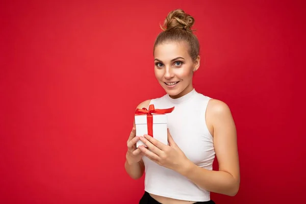Shot of beautiful positive smiling young blonde woman isolated over red background wall wearing white top holding gift box and looking at camera — Stock Photo, Image