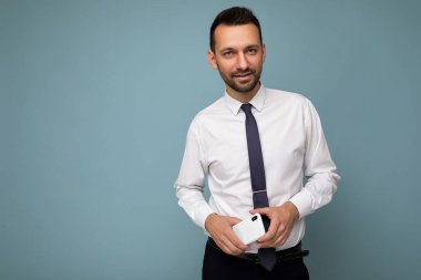 Yakışıklı, esmer, sakallı, beyaz gömlekli ve kravatlı bir adamın fotoğrafı. Mavi arka planda izole edilmiş, el telefonunda boş bir alanda kameraya bakıyor.