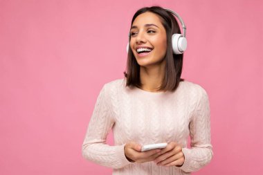 Güzel, neşeli, güler yüzlü, şık kıyafetler giyen, arka plan duvarından izole edilmiş, cep telefonu kullanan, beyaz bluetooth kulaklık takan ve müzik dinleyen genç bir kadının fotoğrafı.