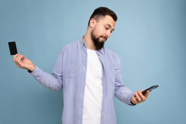 Arkadaki duvarda izole edilmiş günlük giysiler giyen yakışıklı bir adam telefon ve kredi kartı kullanıyor ve akıllı telefon ekranına bakarak ödeme yapıyor.