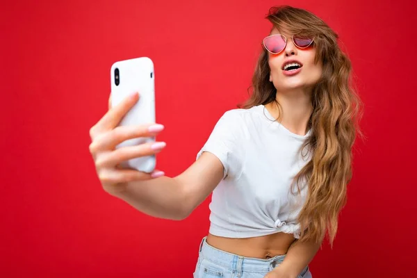 Close-up foto van sexy mooie volwassen blonde vrouw met mobiele telefoon het nemen van selfie foto met behulp van smartphone camera dragen zonnebril dagelijks stijlvolle outfit geïsoleerd over kleurrijke muur achtergrond — Stockfoto