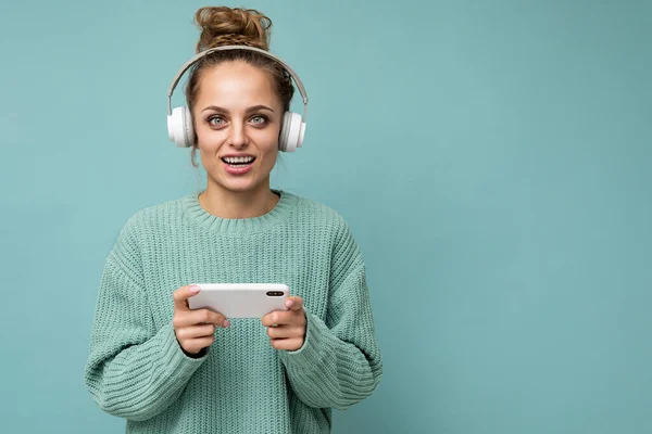 Zbliżenie zdjęcie pięknej pozytywnej młodej kobiety noszącej niebieski sweter odizolowany na niebieskim tle, noszącej białe słuchawki bezprzewodowe bluetooth i słuchającej muzyki oraz korzystającej z telefonu komórkowego — Zdjęcie stockowe