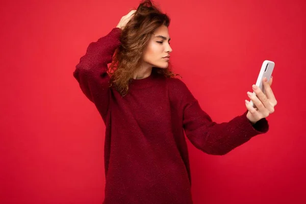 Attractive young blogger woman with curly hair wearing dark red sweater isolated on red background wall holding and using smart phone looking at telephone screen and taking selfie — Stock Photo, Image
