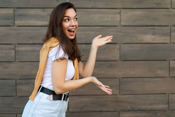 Foto de jovem emocional feliz positivo bela mulher morena vestindo roupas casuais em pé na rua perto da parede e apontando para o espaço de cópia. Conceito de estilo de vida — Fotografia de Stock