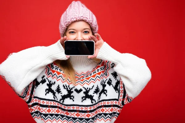 Çekici, mutlu, örgü şapkalı ve kalın kazaklı genç bir kadının fotoğrafı. Kırmızı arka planda izole bir şekilde duruyor. Akıllı telefonlu, boş ekranlı bir kameraya bakıyor.