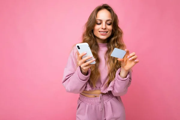 Linda sorrindo positivo jovem loira encaracolado mulher vestindo roupas rosa isolado sobre fundo rosa usando telefone celular pagando compras on-line através de cartão de crédito olhando para exibição de smartphones — Fotografia de Stock