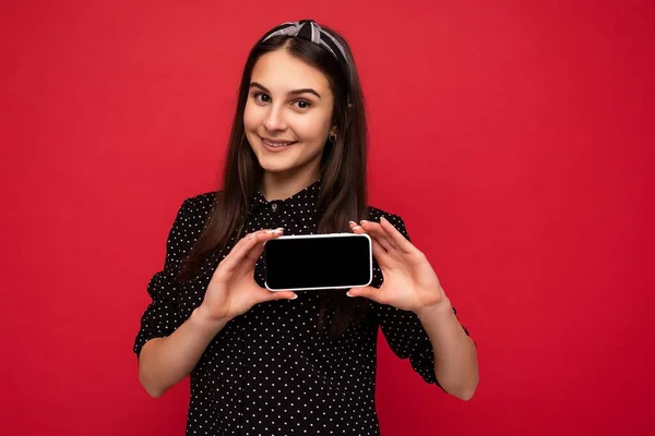 Foto da bela adolescente sorrindo boa aparência vestindo roupa elegante casual em pé isolado no fundo com espaço de cópia segurando smartphone mostrando telefone na mão com tela vazia para mockup — Fotografia de Stock