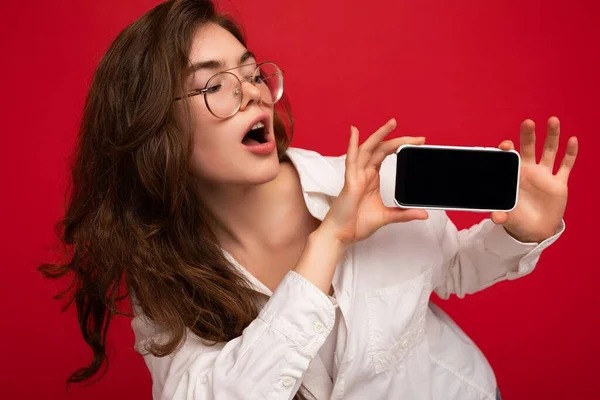 Foto von sexy schockiert schöne junge brünette Frau trägt weißes Hemd und optische Brille isoliert über rotem Hintergrund hält in der Hand und zeigt Handy mit leerem Bildschirm für Ausschnitt suchen — Stockfoto