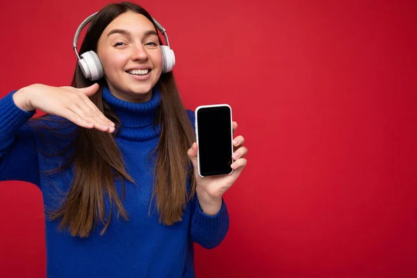Aantrekkelijke vrolijke glimlachende jonge brunette vrouw draagt blauwe trui geïsoleerd over rode achtergrond houden en met behulp van mobiele telefoon surfen op het internet het dragen van witte headsets luisteren naar muziek kijken — Stockfoto