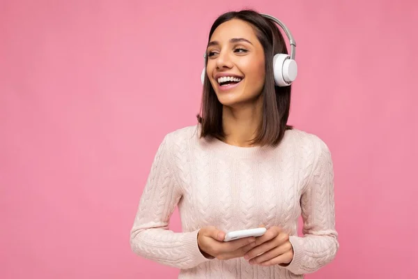 Photo of beautiful joyful smiling young woman wearing stylish casual clothes isolated over background wall holding and using mobile phone wearing white bluetooth headphones listening to music and — Stock Photo, Image