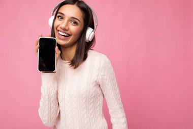 Çekici, pozitif gülümseyen, şık günlük kıyafetler giyen, renkli arka plan duvarında izole edilmiş, cep telefonlu beyaz bluetooth giyimli, boş ekranlı genç bir kadın.