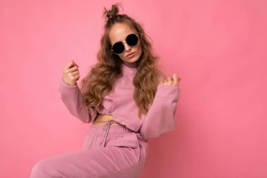 Pembe arka plan duvarında izole edilmiş, pembe spor kıyafetleri ve şık güneş gözlükleri takan, kameraya bakıp dans eden neşeli, genç, esmer, kıvırcık kadın.