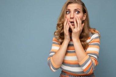 Samimi duyguları olan genç, sarışın, kıvırcık bir kadın çizgili forma giyiyor. Mavi arka planda, boş bir alanda izole edilmiş, korkmuş ve endişeli kadın gözlerini kapatıyor.