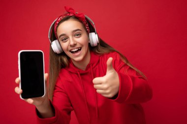 Kırmızı kapüşonlu, neşeli, neşeli, esmer bir kız. Kırmızı arka planda izole edilmiş. Akıllı telefonu beyaz kablosuz kulaklık takıyor ve komik kulaklıklar dinliyor.