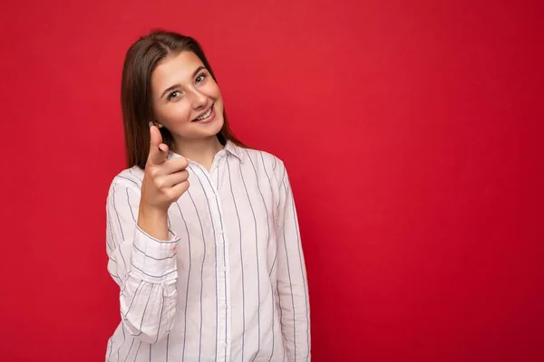 背景色の壁に隔離された誠実な感情を持つ若い笑顔の美しいブルネットの女性は、カジュアルな白いシャツを着てカメラで指を指しています。肯定的な概念 — ストック写真