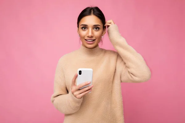 Vonzó aranyos fiatal barna nő visel bézs meleg pulóver áll elszigetelt felett rózsaszín háttér szörfözés az interneten telefonon keresztül nézi a kamera és kérdéseket — Stock Fotó