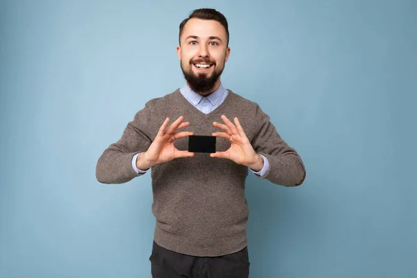 Guapo sonriente morena barbudo hombre con suéter gris y camisa azul aislado en la pared de fondo celebración de la tarjeta de crédito mirando a la cámara — Foto de Stock