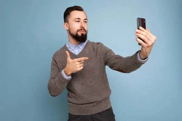 Foto de jovem morena bonito positivo homem sem barba com barba vestindo camisola cinza casual e camisa azul isolada na parede de fundo rosa segurando smartphone tirando foto selfie olhando para celular — Fotografia de Stock