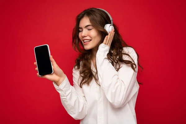매력적 인 긍정적 인 긍정적 인 젊은 갈색 곱슬머리 여성붉은 배경 벽에 고립된 흰색 셔츠를 입고 빈 디스플레이가 장착 된 휴대 전화를 보여 주고 있다. — 스톡 사진