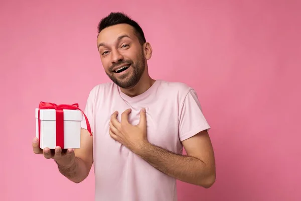 Photo shot of handsome positive surprised brunet young unshaven male person with beard isolated over pink background wall wearing pink t-shirt holding white gift box with red ribbon and looking at — 图库照片