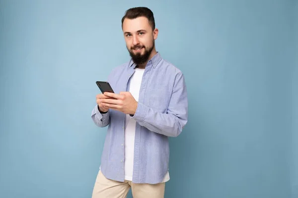 Guapo joven morena sin afeitar hombre con barba usando elegante camiseta blanca y camisa azul aislado sobre fondo azul con espacio vacío sosteniendo en la mano y el uso de sms de mensajería telefónica mirando — Foto de Stock