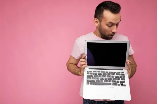 Hombre guapo sorprendido y sorprendido sosteniendo ordenador portátil mirando monitor de computadora en camiseta sobre fondo rosa aislado —  Fotos de Stock