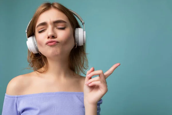Closeup photo of beautiful positive young blonde woman wearing blue crop top isolated over blue background wall wearing white wireless bluetooth earphones listening to cool music and enjoying — Stock Photo, Image
