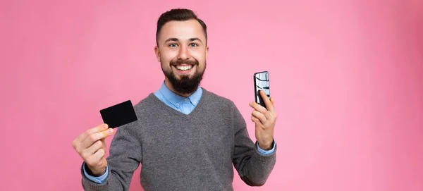 Panoramic photo shot of Handsome smiling brunette bearded young man wearing stylish grey sweater and blue shirt isolated over background wall holding credit card and mobile phone looking at camera — 图库照片