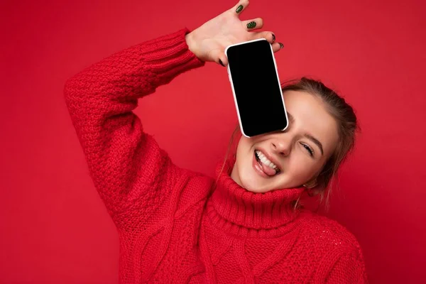 Zdjęcie pięknej uroczej uśmiechniętej młodej kobiety noszącej ciepły czerwony sweter odizolowany na czerwonym tle ściany trzymając smartfona i pokazując telefon z pustym wyświetlaczem do makiety patrząc w aparat — Zdjęcie stockowe