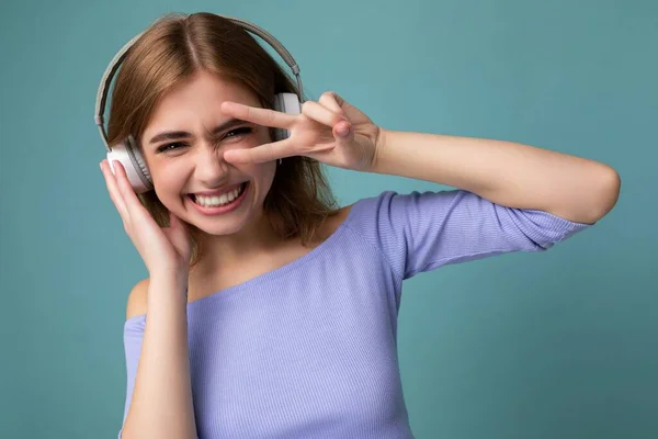 Photo of charming smiling sexy young blonde woman wearing blue crop top isolated on blue background wearing white wireless bluetooth earphones listening to cool music and having fun showing peace — Stock Photo, Image