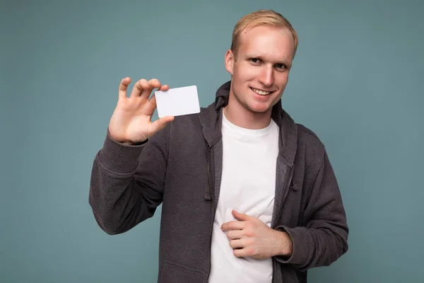 Foto de un guapo hombre rubio positivo usando suéter gris y camiseta blanca aislada sobre una pared de fondo azul sosteniendo la tarjeta de crédito mirando a la cámara —  Fotos de Stock