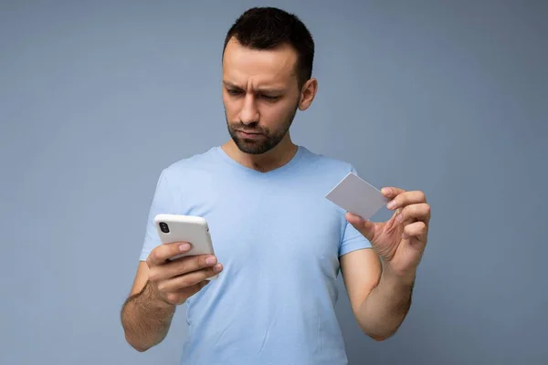 Arkadaki duvarda izole edilmiş günlük giysiler giyen yakışıklı bir adam telefon ve kredi kartı kullanıyor ve akıllı telefon ekranına bakarak ödeme yapıyor. — Stok fotoğraf