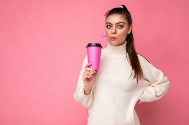 Güzel, genç, pozitif esmer kadın renkli arka plan duvarından izole edilmiş gündelik kıyafetler giyiyor. Makyaj için kağıt bardak tutuyor ve kameraya bakıp kahve içiyor.