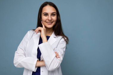 Oldukça çekici, güzel, pozitif gülümseyen bir kadının portresi koyu sarı saçlı, uzun saçlı, mavi arka planda izole edilmiş beyaz bir tıbbi ceket giyen, boş bir alan.