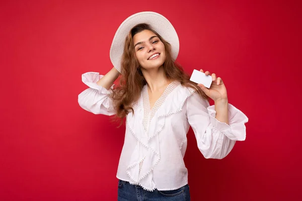 Снимок привлекательного позитивного улыбается молодая темная блондинка женщина в белой блузке и белой шляпе изолированы на красном фоне держа кредитную карту глядя в камеру — стоковое фото