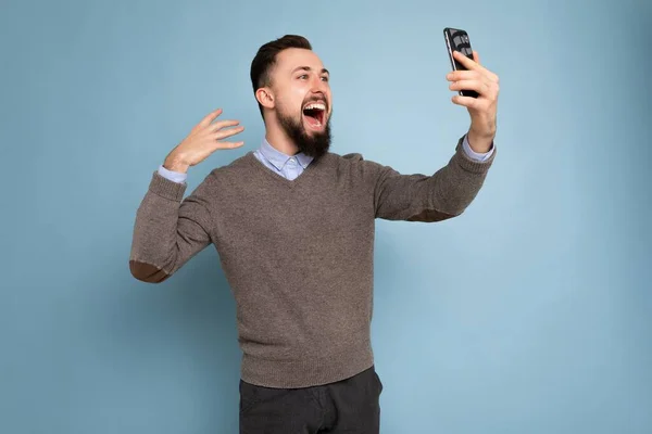 Perfil lateral Foto de joven morena guapo positivo sin afeitar hombre con barba usando suéter gris casual y camisa azul aislado en la pared de fondo rosa celebración de teléfono inteligente que tiene aspecto de videollamada —  Fotos de Stock