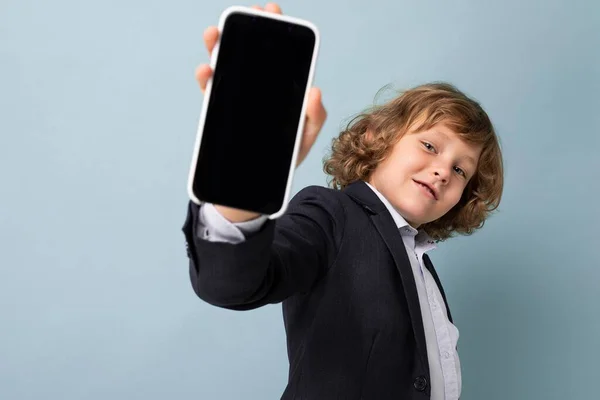 Guapo niño sonriente positivo con pelo rizado usando traje sosteniendo teléfono aislado sobre fondo azul mirando a la cámara y mostrando teléfono inteligente con pantalla de visualización vacía — Foto de Stock