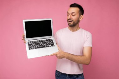 Yakışıklı, şaşkın ve mutlu esmer adam dizüstü bilgisayarını tutuyor. İzole edilmiş pembe arka plandaki netbook monitörüne bakıyor.