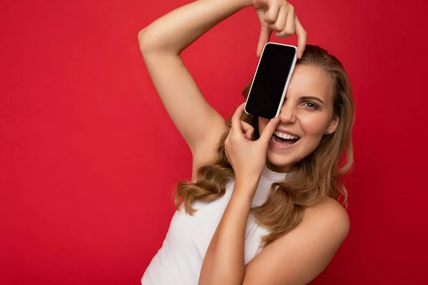 흰색 티셔츠를 입고 빨간 배경에 고립된 금발의 젊은 여성 이 스마트폰을 손에 들고 카메라를 보기 위해 빈 화면을 보여 주는 스마트폰을 들고 있는 사진. — 스톡 사진