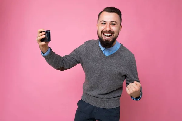 매일 회색 스웨터를 입고 파란 셔츠를 입은 행복 한 젊고 잘생긴 갈색 머리의 남자가 백그라운드 벽에 고립되어 스마트폰으로 비디오를 보고 카메라와 카메라를 보고 있다. — 스톡 사진