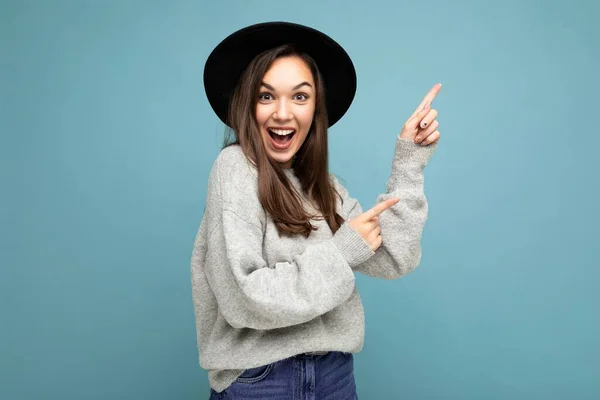 Foto de morena bonita positiva surpreendeu jovem dirigindo os dedos para o lado com espaço vazio, demonstrando apresentar cartaz de preços baixos, vestindo camisola cinza e chapéu preto, isolado em azul — Fotografia de Stock