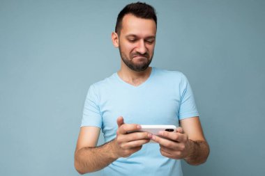 Her gün mavi tişört giyen yakışıklı genç adama mavi arka planda izole edilmiş cep telefonu iletişimini internetten gadjet ekranına bakarak sormanın fotoğrafı.