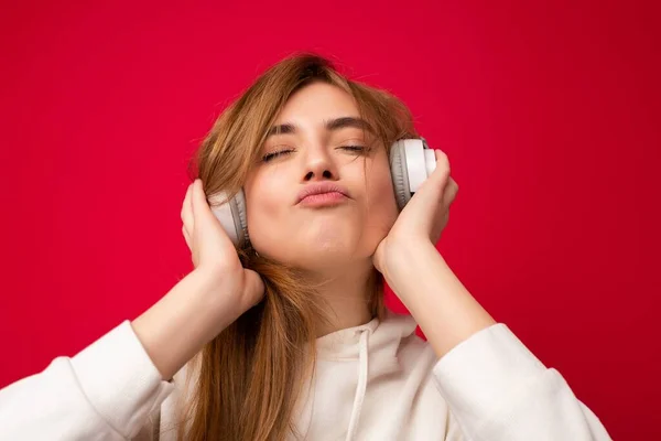 Portrait photo shot of attractive emotional positive young blonde woman wearing white hoodie isolated over colourful background wearing white wireless bluetooth headsets listening to music and — Stock Photo, Image