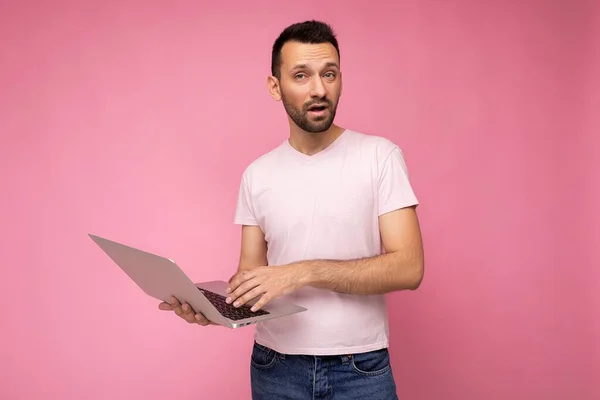 휴대용 컴퓨터를 들고 있는 브루 넷 남성에게 물어 보며 핑크 색 배경으로 티셔츠를 입고 카메라를 보고 있는 손을 보여 주는 모습 — 스톡 사진