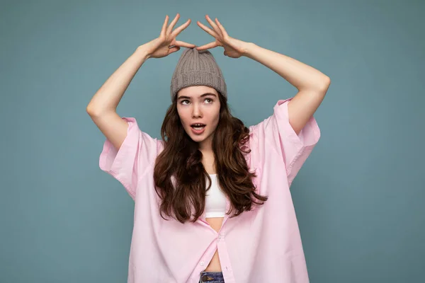 Foto der schönen emotionalen jungen brünetten Frau, die isoliert auf blauem Hintergrund Wand trägt rosa Hemd und grauen Hut, der zur Seite schaut — Stockfoto