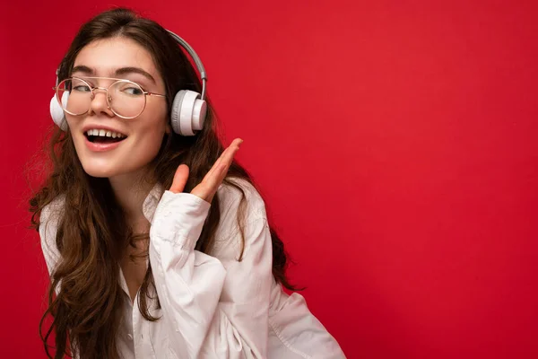 Photo of beautiful happy smiling young brunette woman wearing white shirt and optical glasses isolated over red background wall wearing white wireless bluetooth headphones listening to cool music — Stock Photo, Image