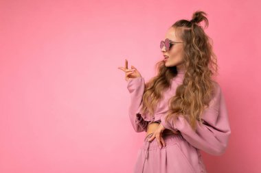 Güzel, genç, üzgün, esmer, kıvırcık saçlı bir kadının pembe arka plan duvarından izole edilmiş, günlük pembe spor kıyafetleri ve şık güneş gözlüğü takıp yan tarafa bakıp sorular sorduğu bir kare.