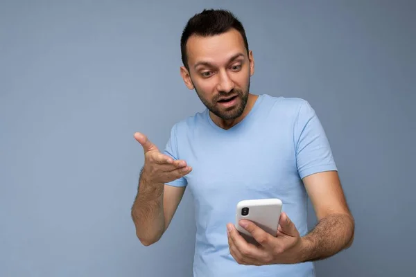 Photo of handsome young man with beard wearing everyday blue t-shirt isolated over blue background holding and using mobile phone communication online on the internet looking at gadjet display and don — Stock Photo, Image