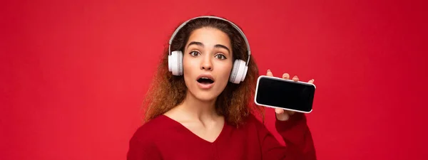 Foto panorâmica de atraente surpreendido jovem morena encaracolado mulher vestindo camisola vermelha escura isolada no fundo vermelho vestindo fones de ouvido brancos ouvindo música e mostrando telefone moderno com vazio — Fotografia de Stock